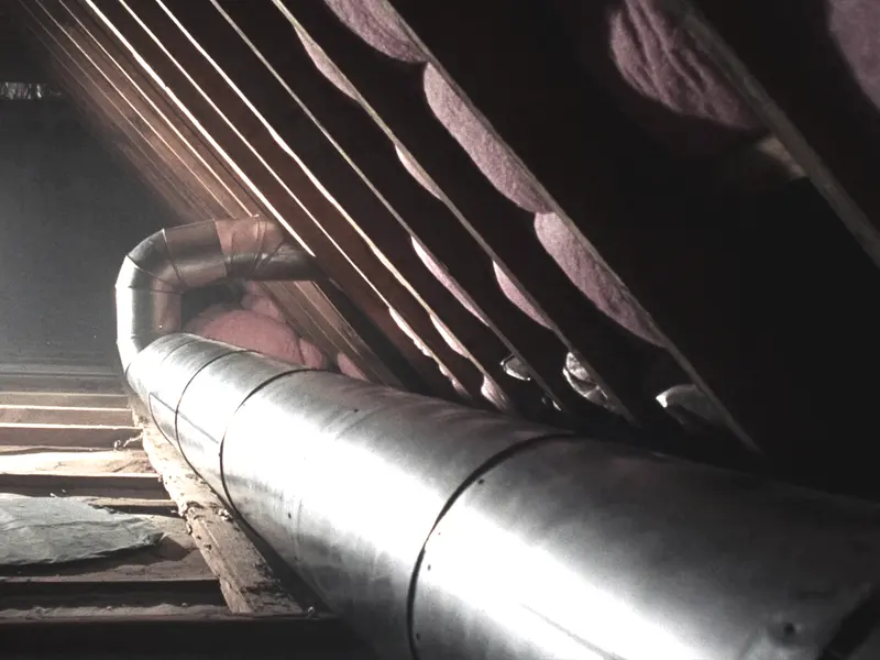 A run of new galvanized round duct between attic rafters, pink insulation batts around it and a work light in the corner