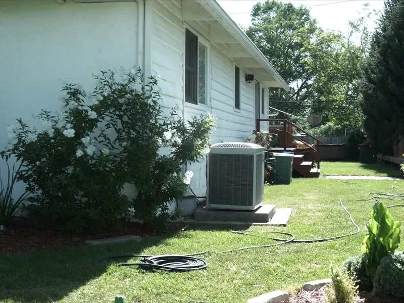 An air conditioning condenser unit on a pad beside white siding, with hostas, a flowering shrub and a garden hose on the lawn