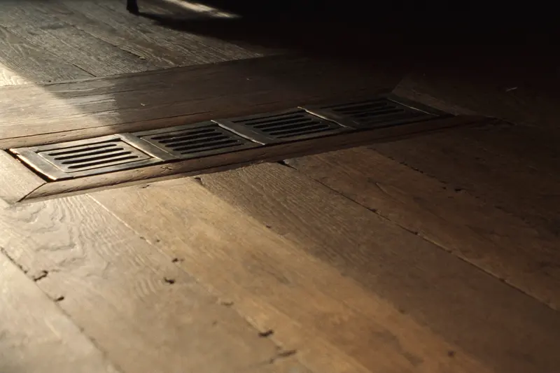 A bronze floor register set into old oak floorboards, lit by low raking light