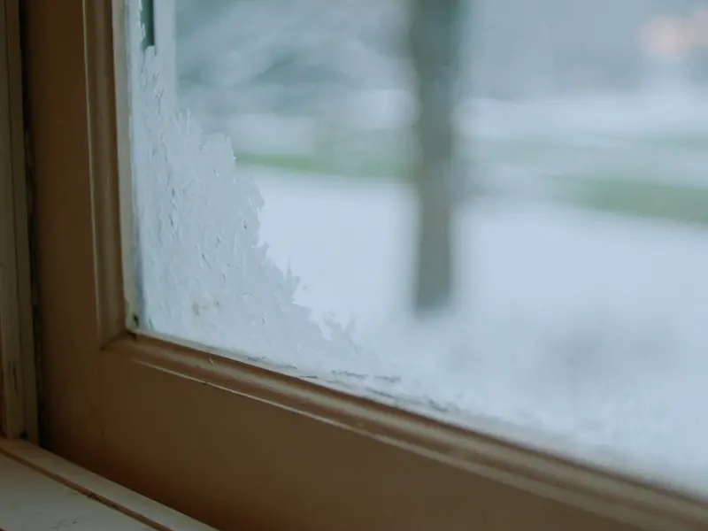 Frost crystals on the corner of an old wooden window at dawn, a snowy yard blurred beyond the glass