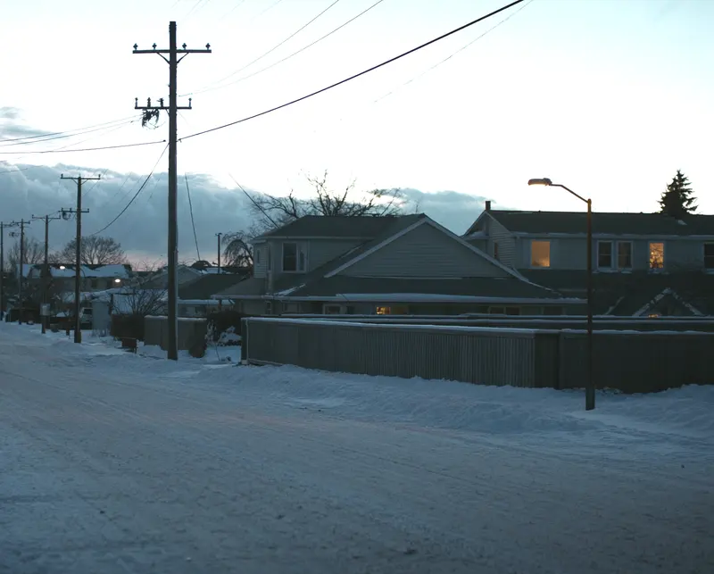 A snow covered residential street at first light, utility poles along the road and warm light in the windows of the houses