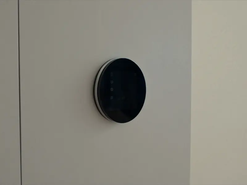 A round dark thermostat with a metal ring mounted on a pale wall, lit from the side