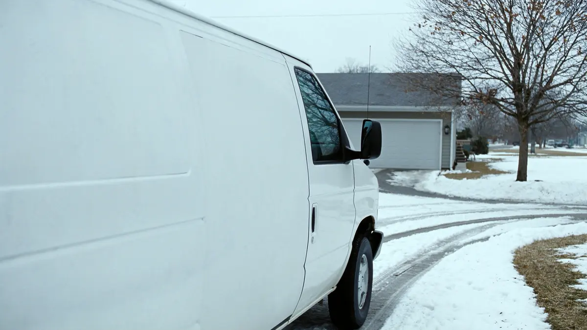 The blank white side of a work van parked in a snowy suburban driveway, with a garage and a bare maple tree behind it