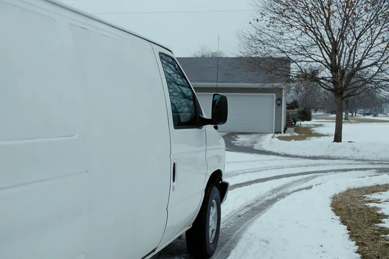 The blank white side of a work van parked in a snowy suburban driveway, with a garage and a bare maple tree behind it